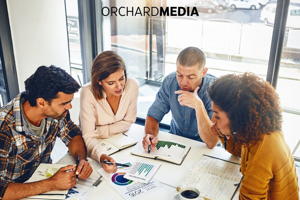 Four diverse professionals gathered around a table analyzing performance charts and graphs during a strategic meeting, with the Orchard Media logo displayed above.
